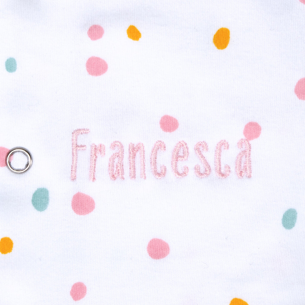 Close up of  the name "Francesca" in pink thread embroidered on a Baby grow with neutral  dotty pattern design with orange trim.